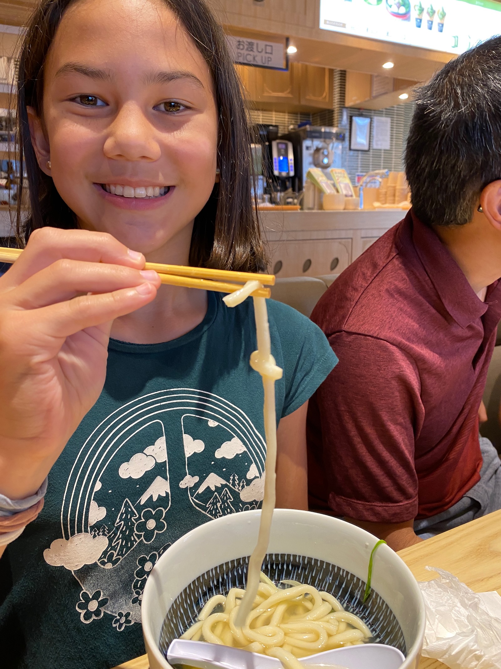 A girl in a green shirt uses chopsticks to hold up an udon noodle with a knot in it.