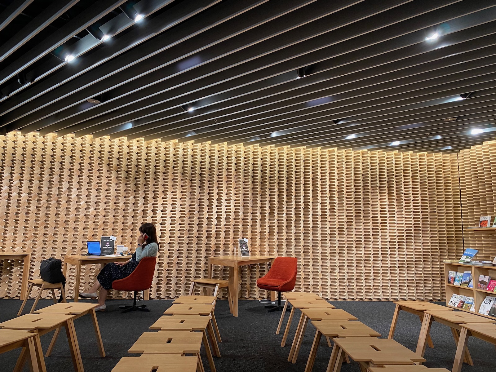 A large room with a patterned wooden wall. Small wooden stools are arranged in rows in the foreground, while in the background a woman seated at a small table speaks on her cellphone.